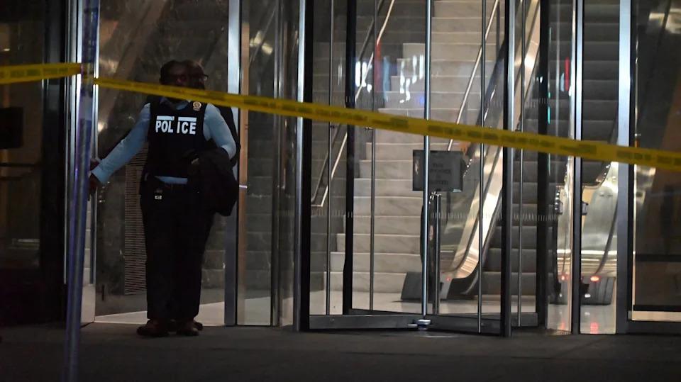 A police officer stands by glass doors with yellow caution tape across the entrance. An escalator is visible inside.