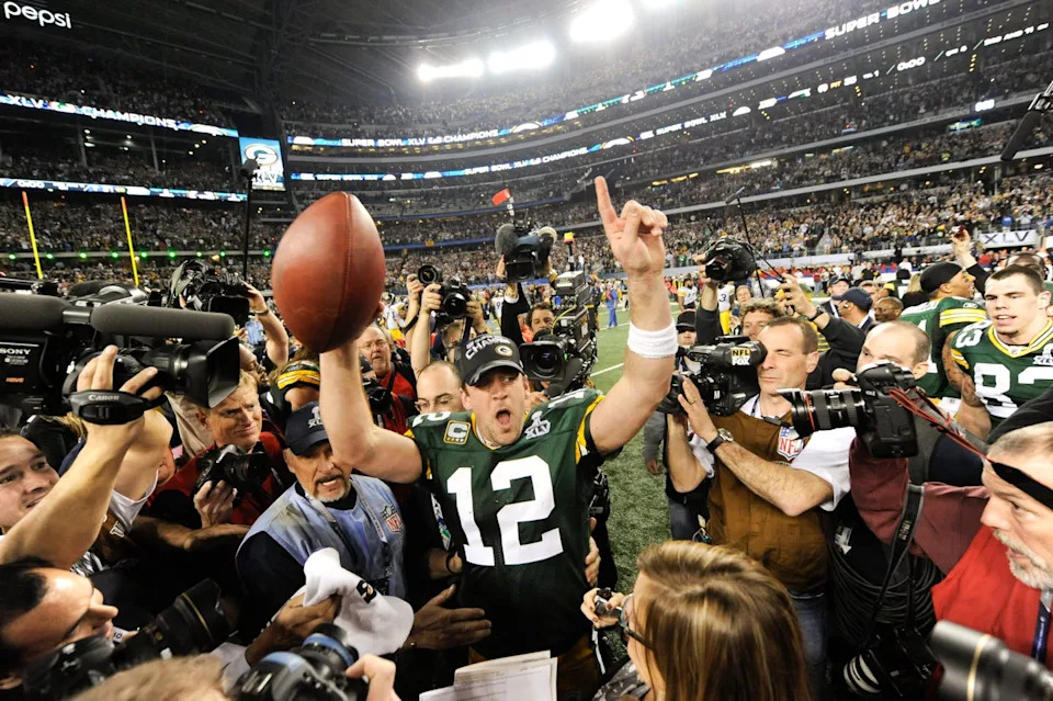 aaron rodgers stands in a crowded football stadium and is surrounded by photographers as he raises a football and one finger in the air in victory, he wears a green bay packer jersey and black hat