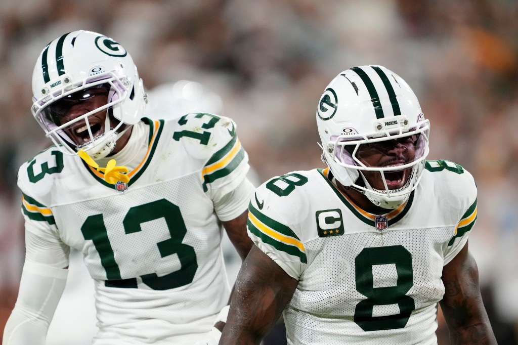 Two Green Bay Packers players, wearing white jerseys with green and yellow stripes and white helmets, celebrate on the field.