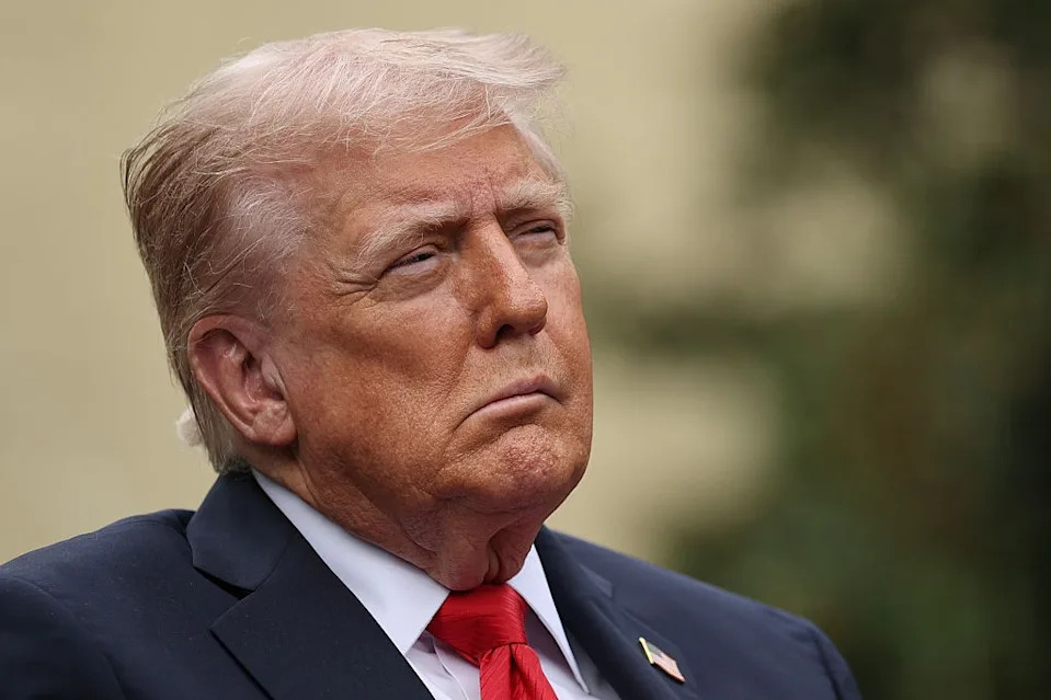 A person in a suit and red tie looks to the side with a serious expression against a blurred background