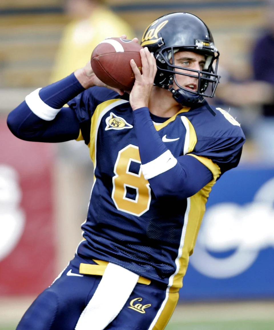 aaron rodgers looking downfield while gripping a football, he wears a helmet and blue and gold uniform with a white towel at his waist