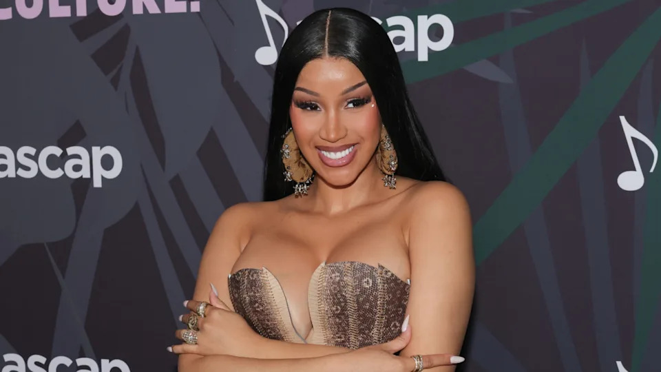 Cardi B poses confidently with crossed arms, wearing a strapless dress and large earrings, against a backdrop with musical notes.