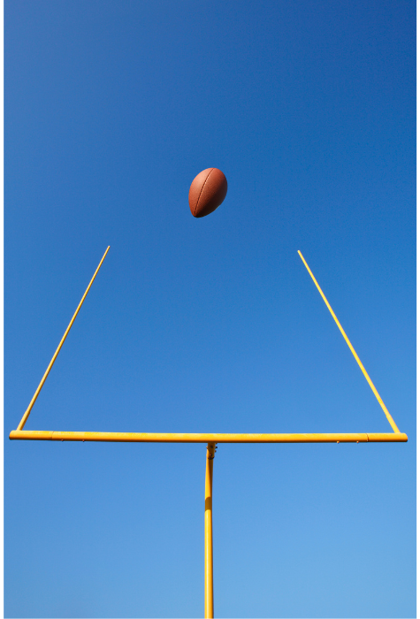 Looking up at a yellow field goal against a perfectly blue sky with a football flying between the poles.