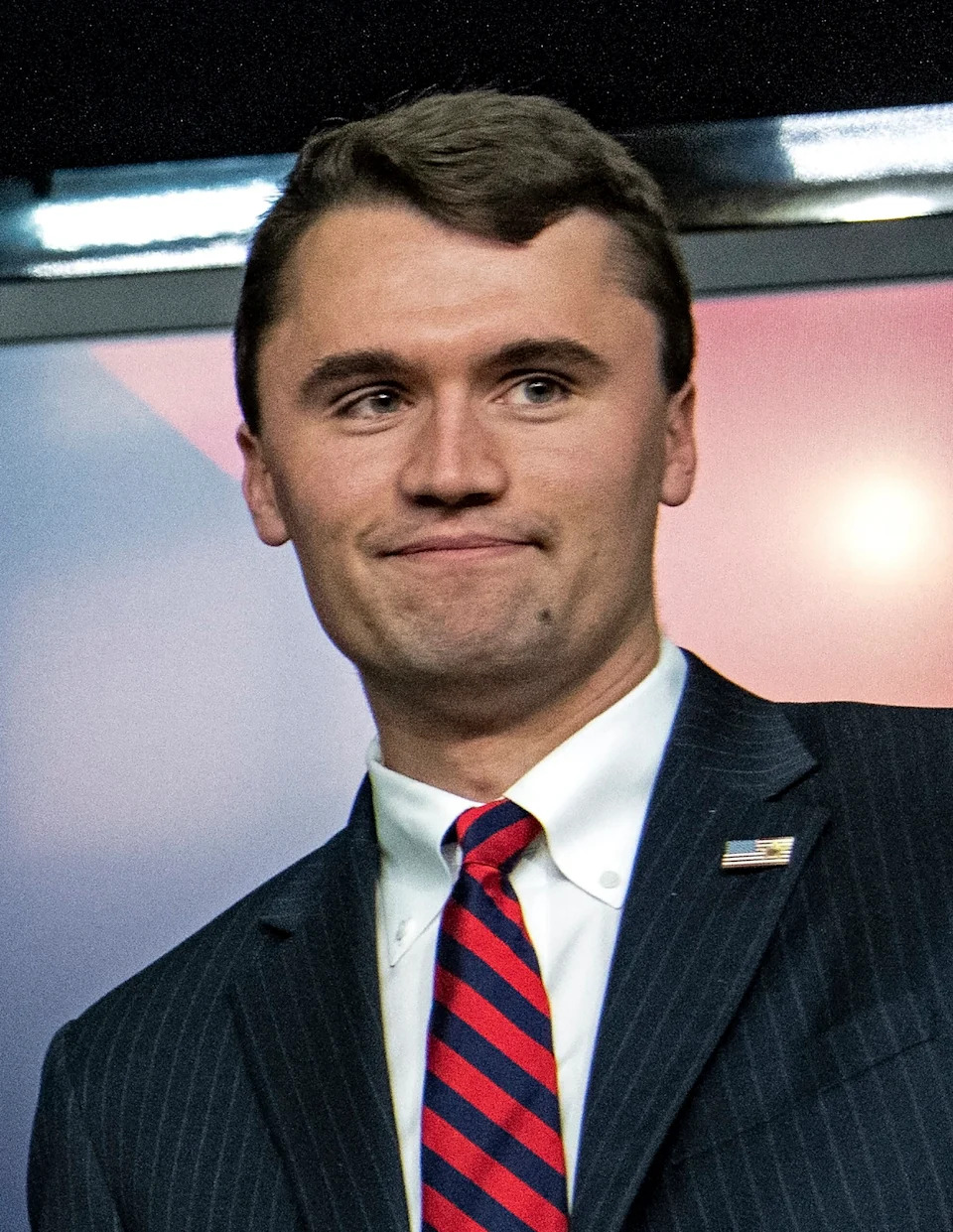 Charlie Kirk in a dark pinstripe suit with a red and blue striped tie, looking off to the side during a public appearance.