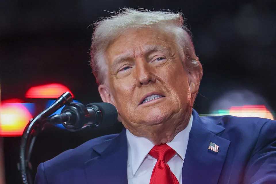 A person in a suit and red tie speaks at a podium, looking to the side. There are blurred lights in the background