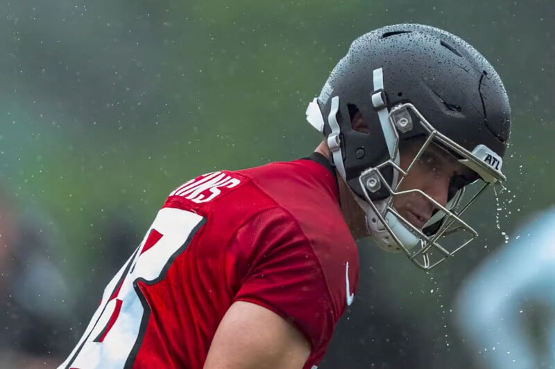 A football player wearing a black helmet and red jersey, with rain droplets visible on his helmet and shoulder, looks to the side during a rainy outdoor practice or game.