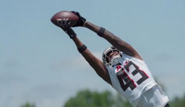 A football player in a white jersey with the number 43 stretches his arms overhead to catch a football during a practice, with a blue sky and trees in the background.