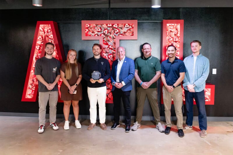 Seven people stand in a row smiling in front of large red letters spelling "ATL" on a black wall. One person holds a trophy or award. They are casually and semi-formally dressed.