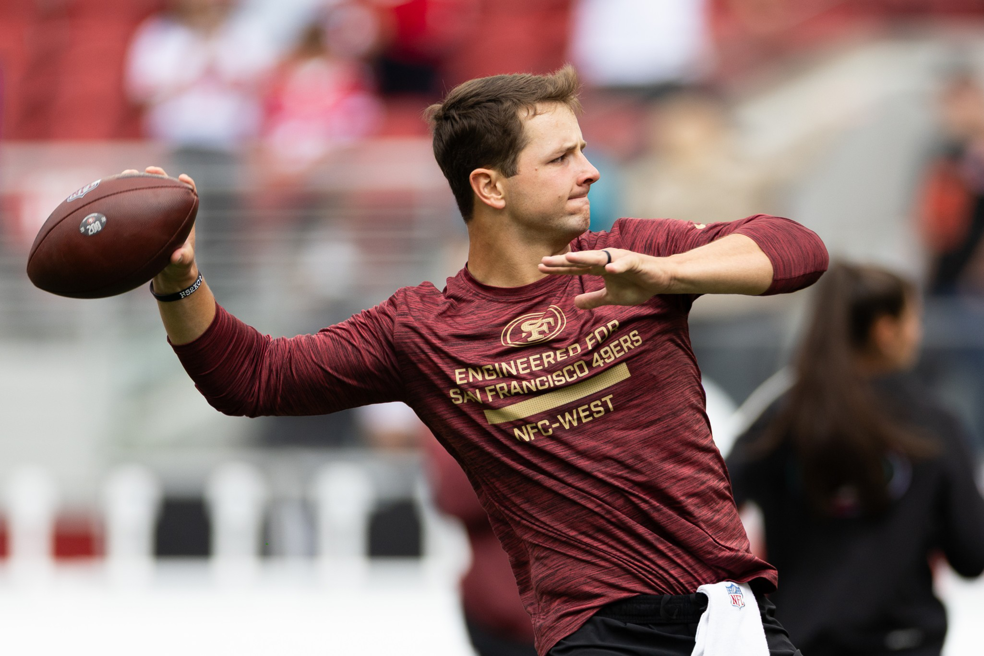 A man wearing a maroon San Francisco 49ers shirt prepares to throw a football during what appears to be a warm-up or practice session.
