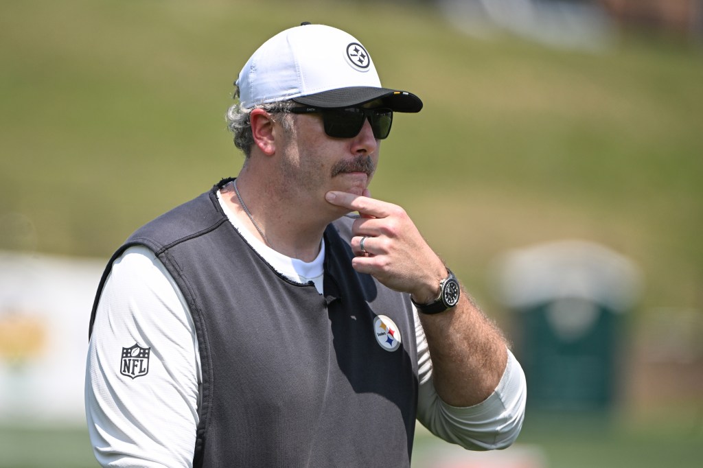 Pittsburgh Steelers coach at training camp, wearing a white hat, black sunglasses, and a black vest, with his hand on his chin in thought.