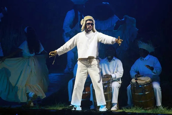 Performer in white outfit and hat stands on stage gesturing outward, surrounded by seated drummers in white
