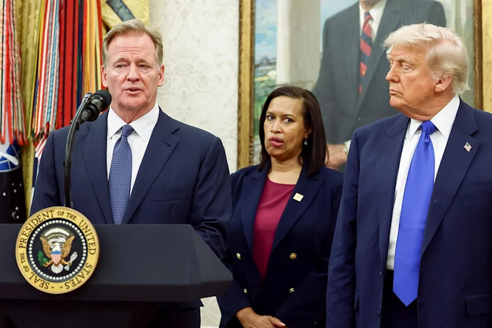 Three people stand at a podium featuring the presidential seal, wearing formal suits. The man at the podium speaks, while the others look on