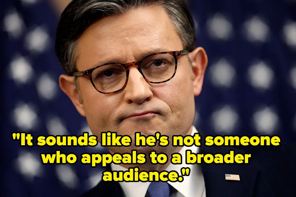 A person in a suit and tie stands against a patterned backdrop, looking contemplative with glasses on
