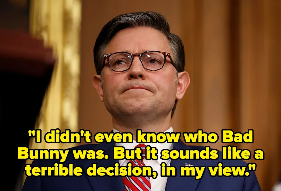 A person in a formal suit with glasses stands in a professional setting, appearing thoughtful