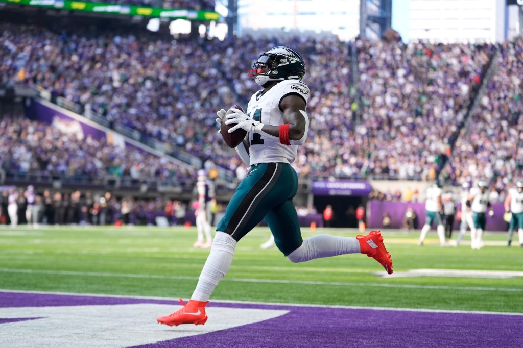 An Eagles player in a white and teal uniform with number 14 on the chest, wearing bright red cleats, runs across the football field holding a football.