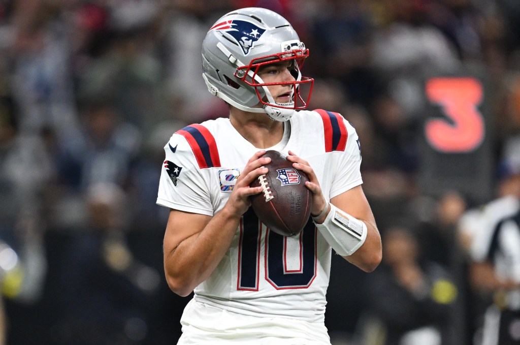 New England Patriots quarterback Mac Jones in his team's white jersey and silver helmet, holding a football.