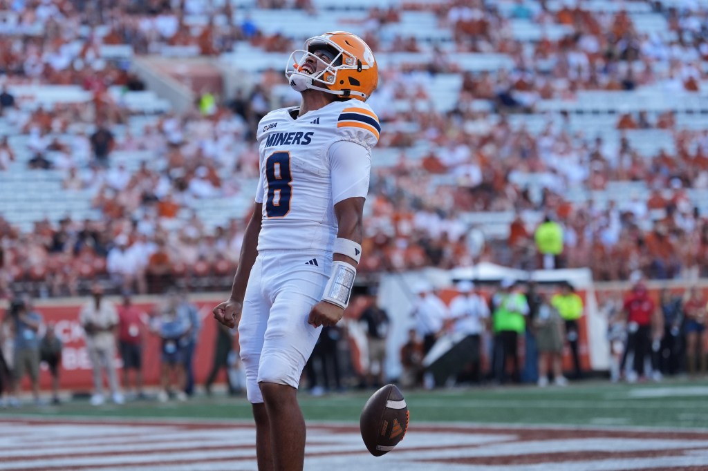 A UTEP football player in a white uniform with "Miners" and number 8, wearing an orange helmet, looks up with a football floating near his waist.