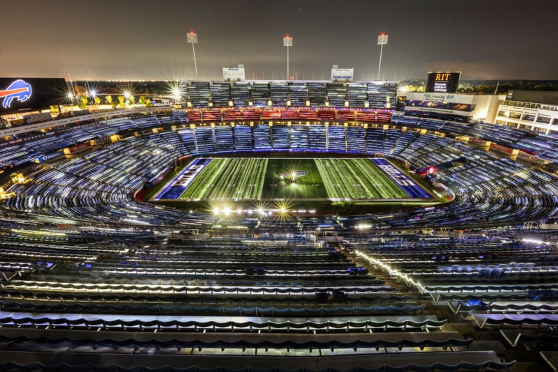 A brightly lit football stadium at night, with empty seats and the field illuminated. The Buffalo Bills logo is visible at midfield, and various advertisements are displayed around the stadium.