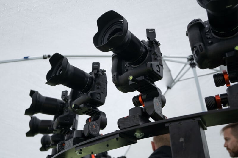 Several professional cameras mounted on a metal rig under a white canopy, viewed from below. Two people with short hair are partially visible beneath the equipment.