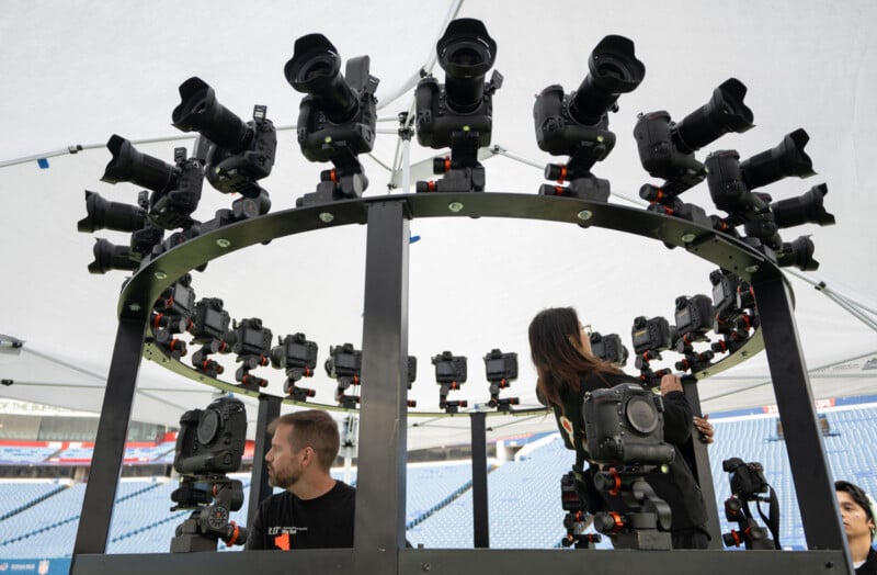 A man and a woman stand inside a circular rig equipped with numerous professional cameras, arranged in multiple rows, under a large tent at a stadium.