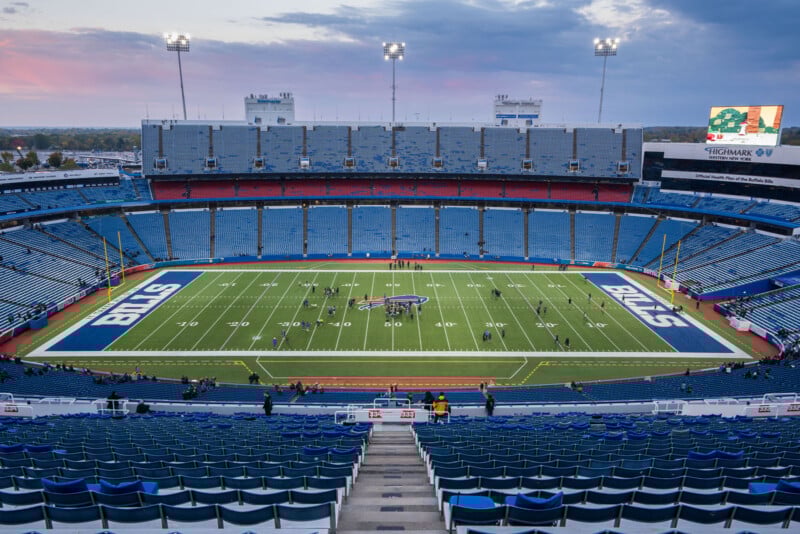 Wide view of an empty football stadium with "BILLS" in the end zones, a few people on the field, blue seats throughout, and a digital scoreboard on the right side against a cloudy sky at sunset.