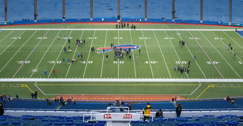 A football field with a buffalo logo at the center, scattered people walking on the turf, and mostly empty blue stadium seats surrounding the field.