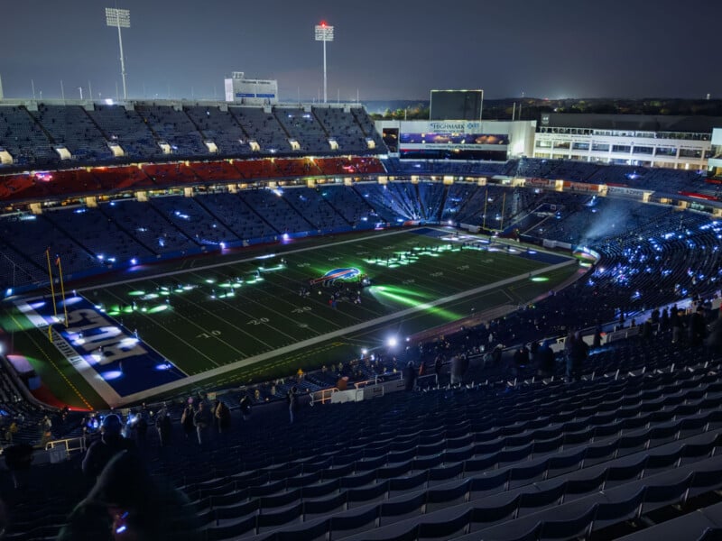 A football stadium at night with bright lights and projections on the field. The seats are mostly empty, and a colorful logo is illuminated at the center of the field. The atmosphere is calm and quiet.