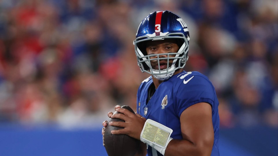 Russell Wilson in a blue Giants uniform and helmet, holding a football.