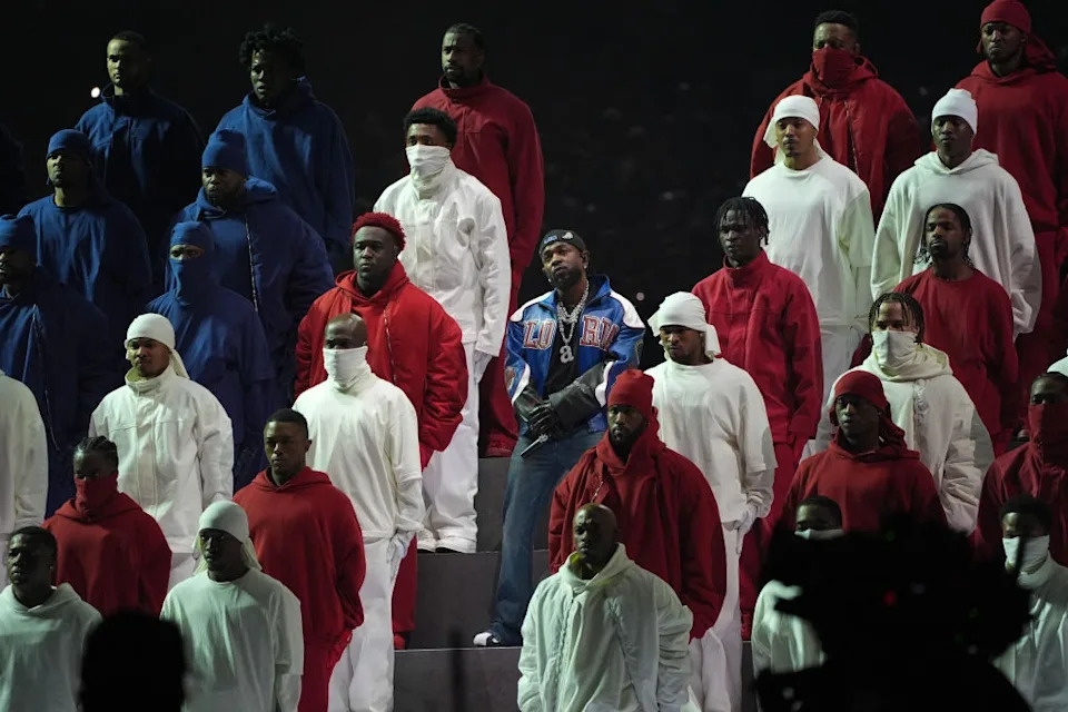 A large group of performers in red, white, and blue streetwear stand on stage in a tiered formation. One central figure wears an orange and blue jacket