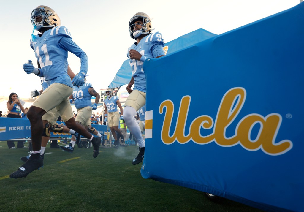 UCLA Bruins football players run onto the field from a tunnel with blue UCLA branding.