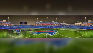 A wide-angle view of an empty football stadium at night, with bright lights and a large buffalo logo at midfield. A few people stand on the turf near the center. Blue seats fill the stands in the background.