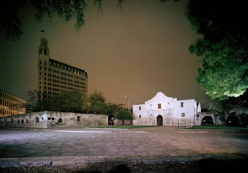 The Alamo at night in San Antonio, Texas, illuminated with a dark sky overhead and surrounding trees and historic buildings in the background.
