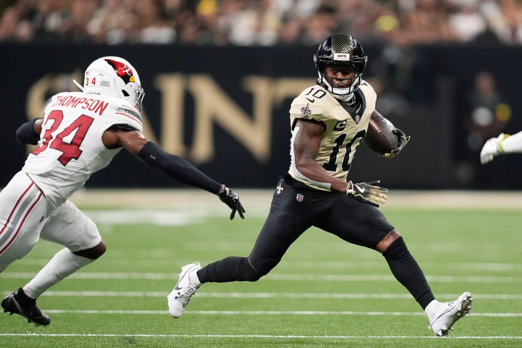 New Orleans Saints player with number 10, wearing black and gold uniform and helmet, running with a football as a player from the Arizona Cardinals wearing white and red uniform with number 34 chases him.
