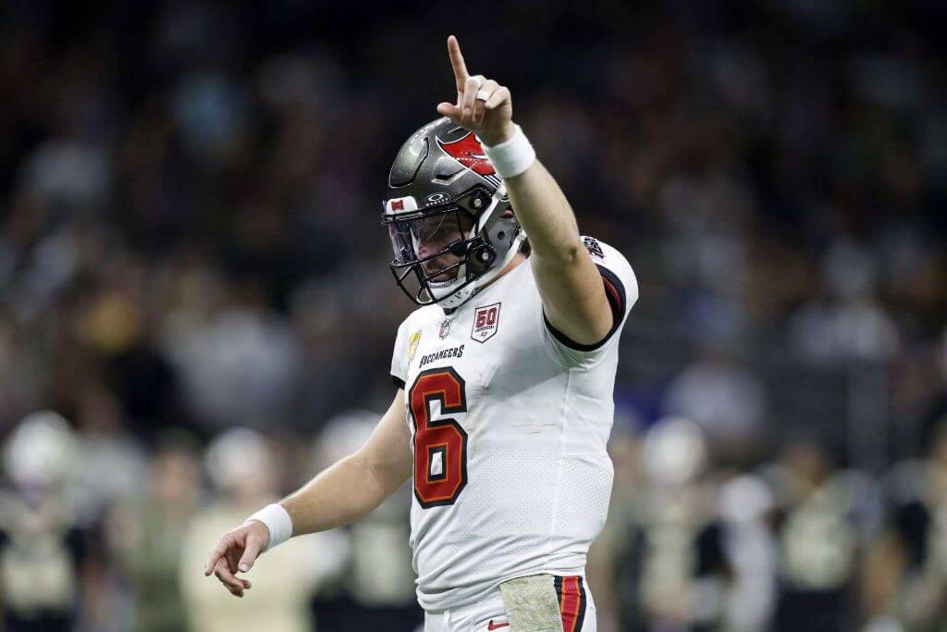 Baker Mayfield is shown from the waist up and from the side, wearing a white Tampa Bay jersey and champagne helmet. His right hand is raised, with his index finger extended.