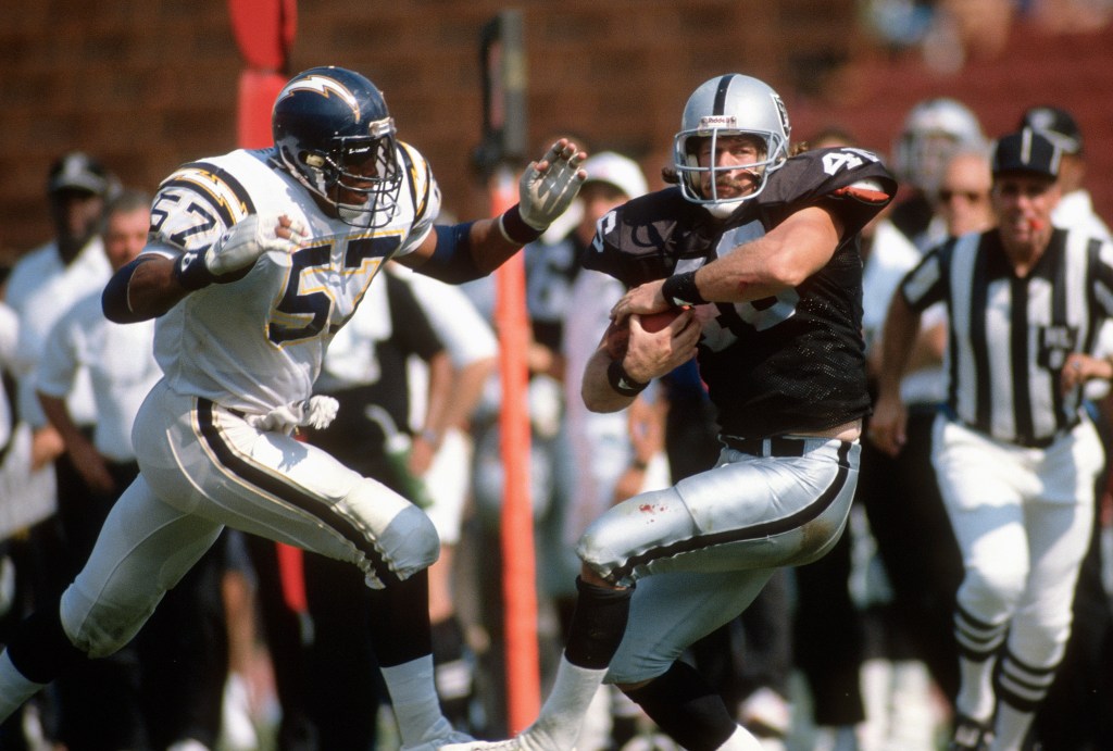 Two football players, one in a white San Diego Chargers uniform and one in a black Los Angeles Raiders uniform, facing each other on the field with a referee in the background.