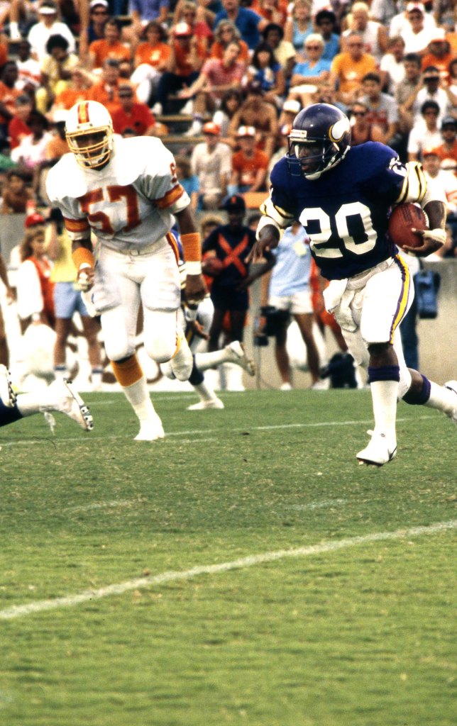 A Minnesota Vikings player in a purple jersey with the number 20 running with the football, pursued by a Tampa Bay Buccaneers player in a white jersey with the number 57.