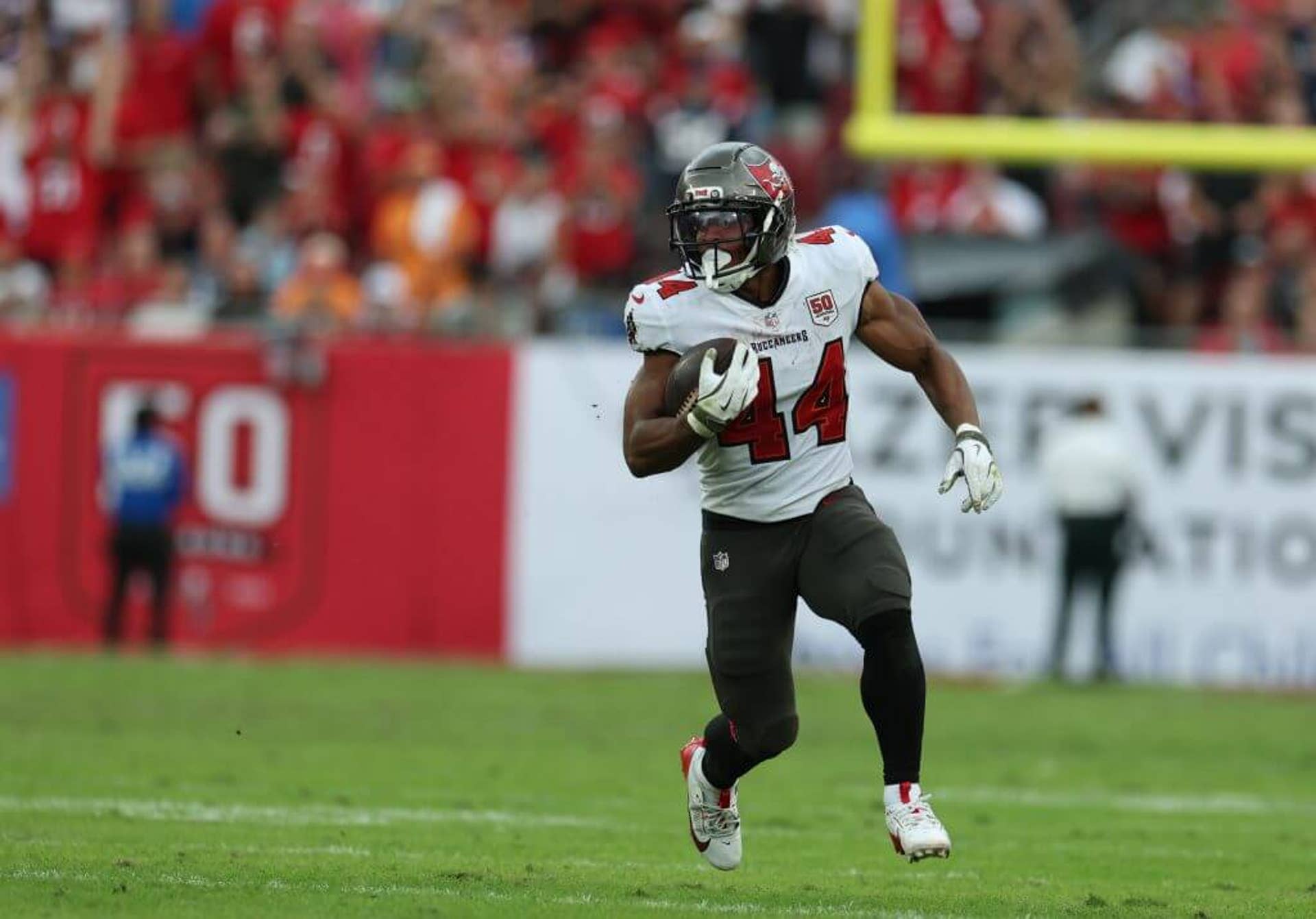 Tampa Bay running back Sean Tucker, wearing a white Buccaneers jersey with pewter-colored pants and helmet, strides in the open field with the ball tucked into his right side.