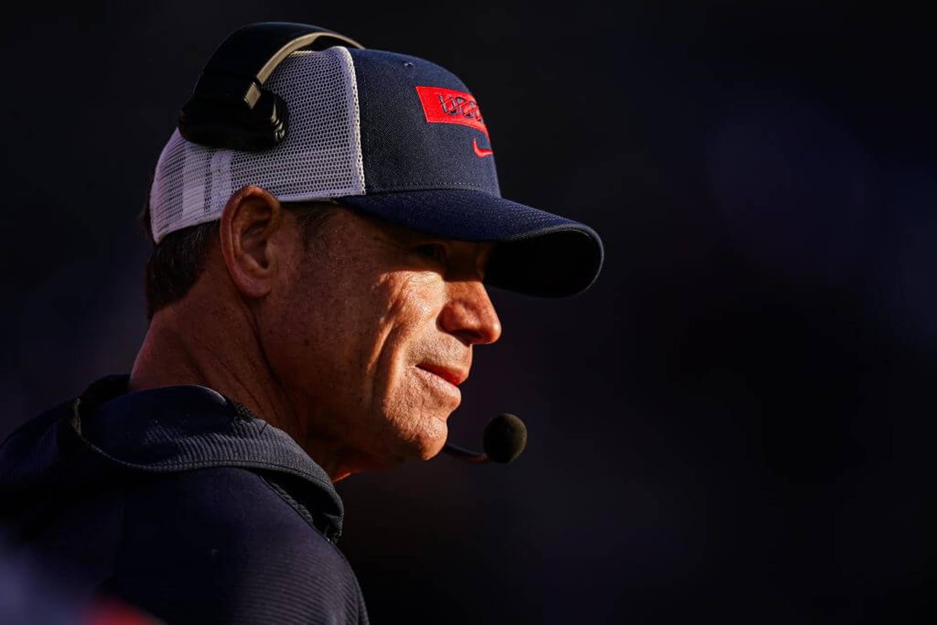 UConn coach Jim Mora, in a ball cap and headset, watches from the sideline during a game.