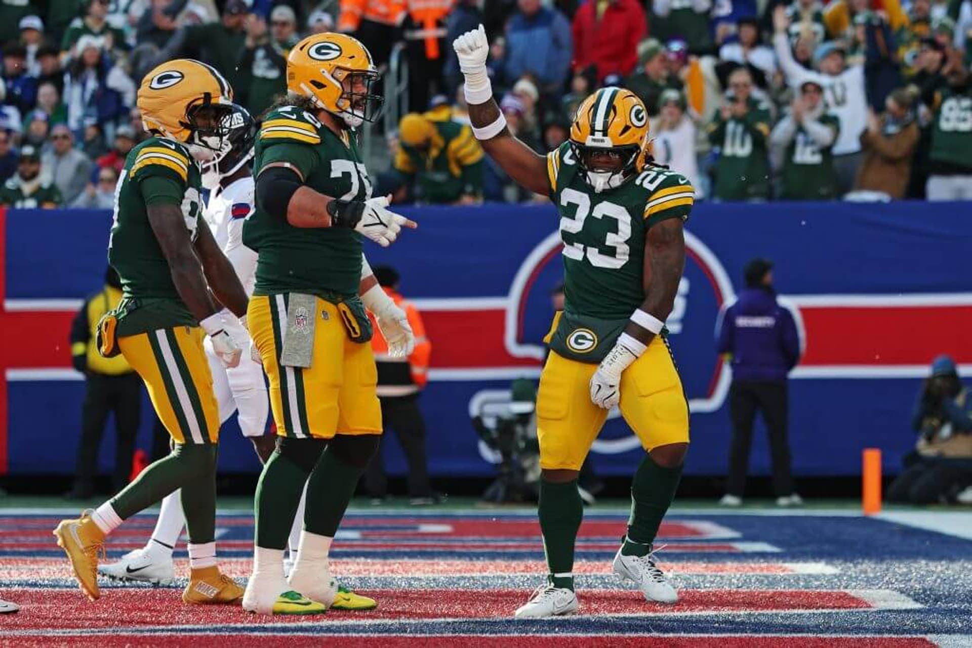 Green Bay running back Emanuel Wilson, wearing a green Packers jersey with yellow pants and helmet, dances with right arm raised in the air after scoring a touchdown while his teammates look on.