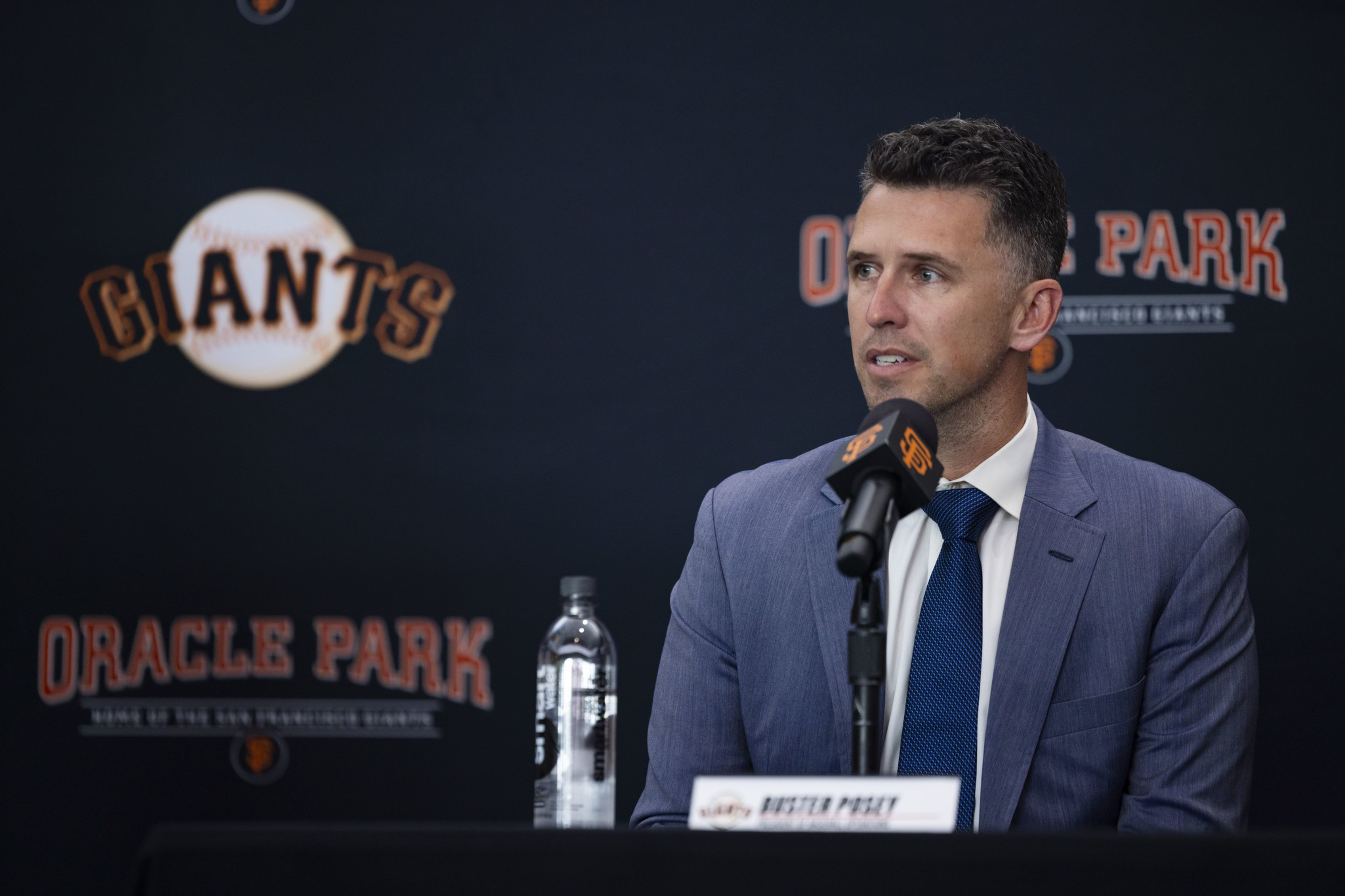 A man in a blue suit and tie speaks at a press conference with a Giants logo and Oracle Park backdrop behind him.