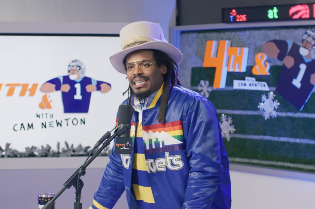 Cam Newton in a blue jacket with a Denver Nuggets logo and a white hat, speaking into a microphone.