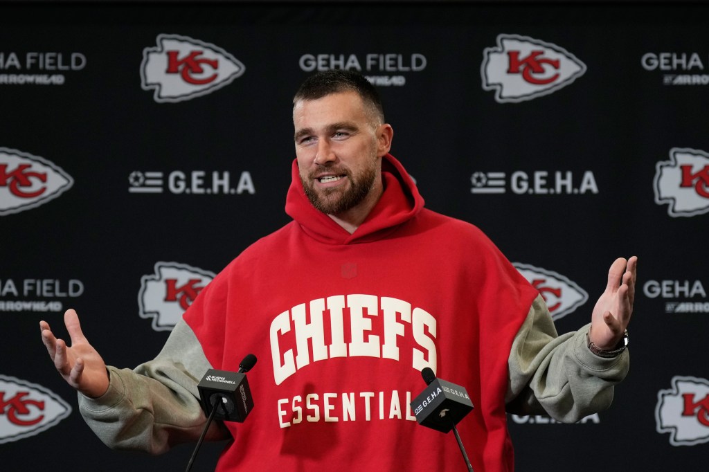 Travis Kelce speaking at a press conference with a Chiefs Essential hoodie.