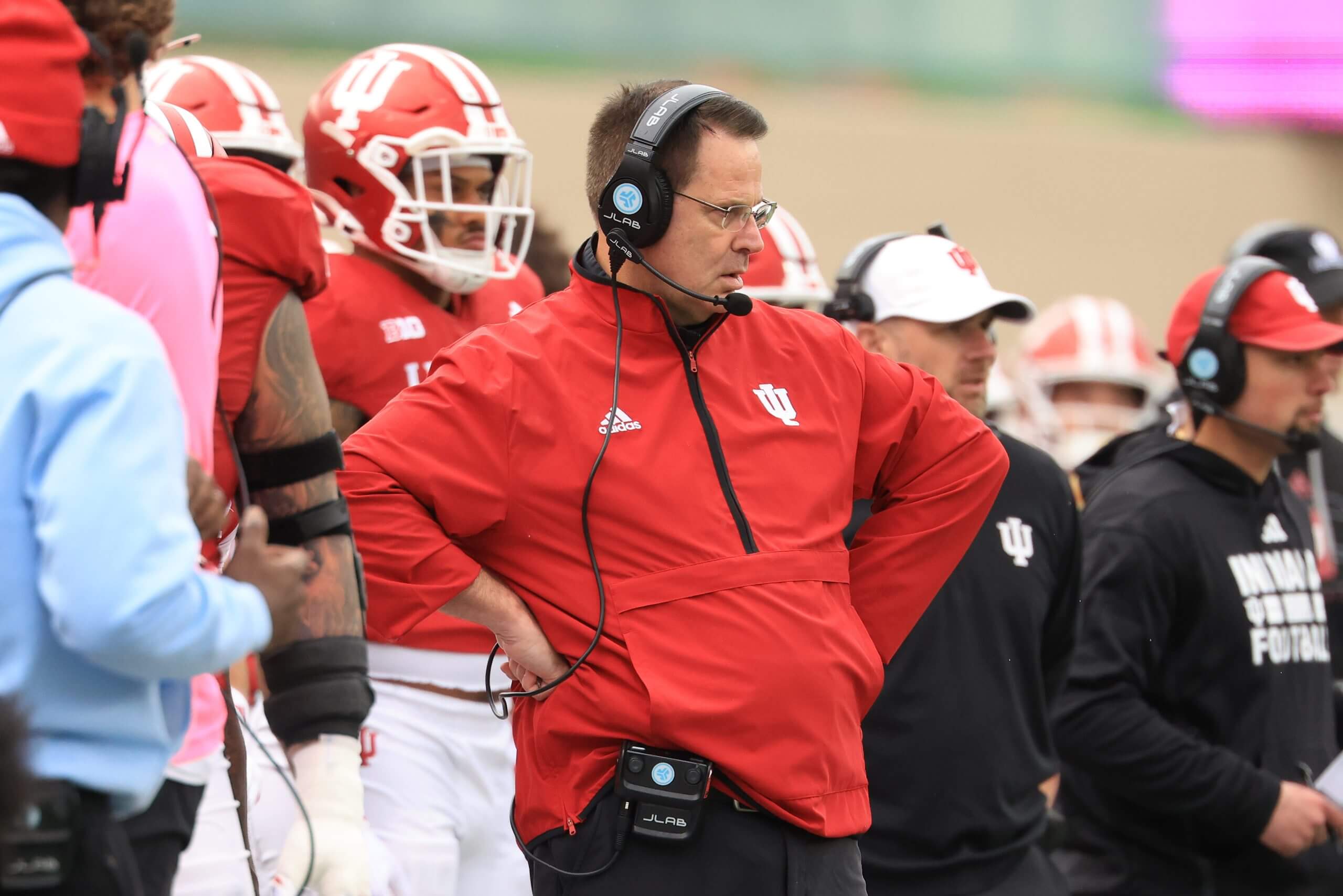Curt Cignetti on the sideline in a red Indiana pullover, hands on his hips, wearing glasses and a headset.
