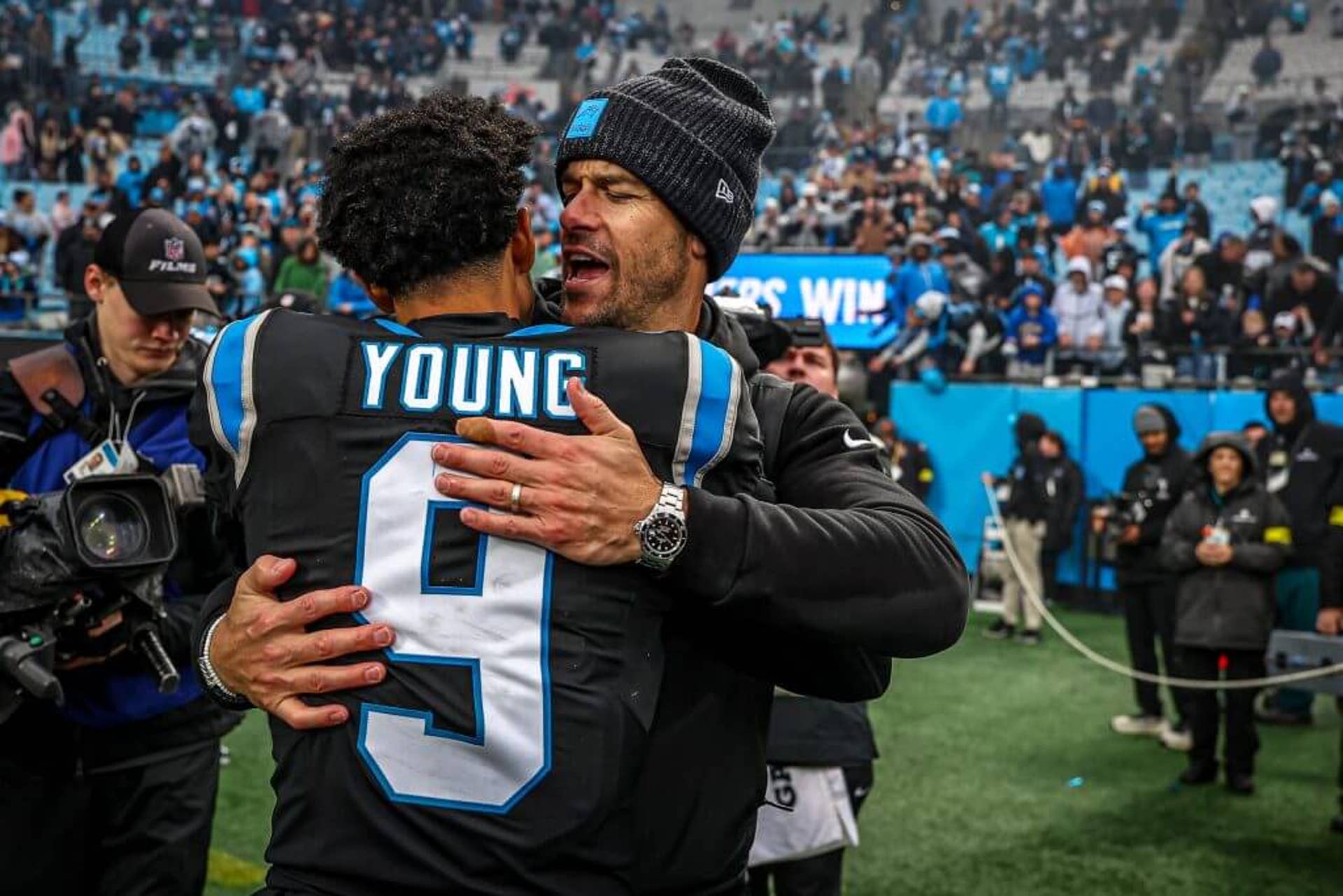 Carolina Panthers Coach Dave Canales hugs quarterback Bryce Young on the field postgame as Panthers fans cheer in the background.