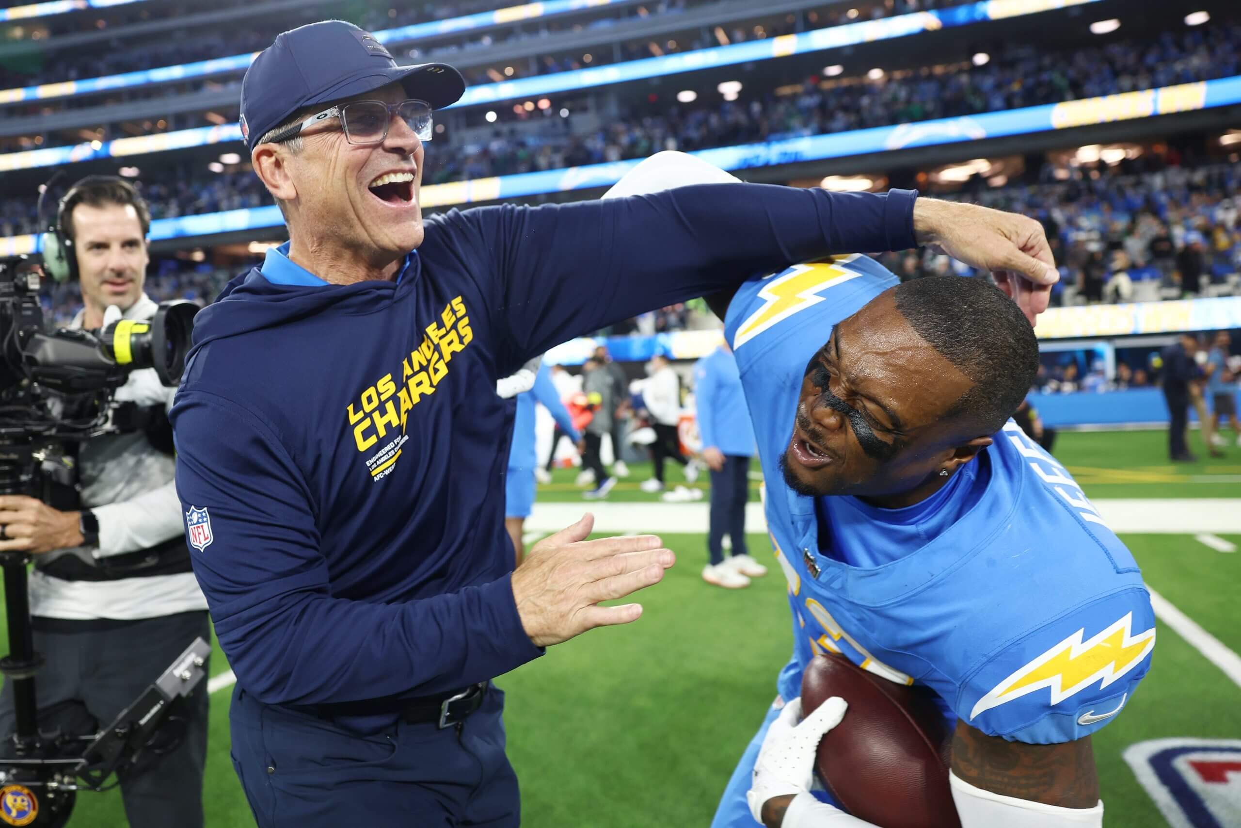 A smiling coach Jim Harbaugh, left, in a hoodie and ball cap, puts his arm around Tony Jefferson, right, holding the ball on the field.