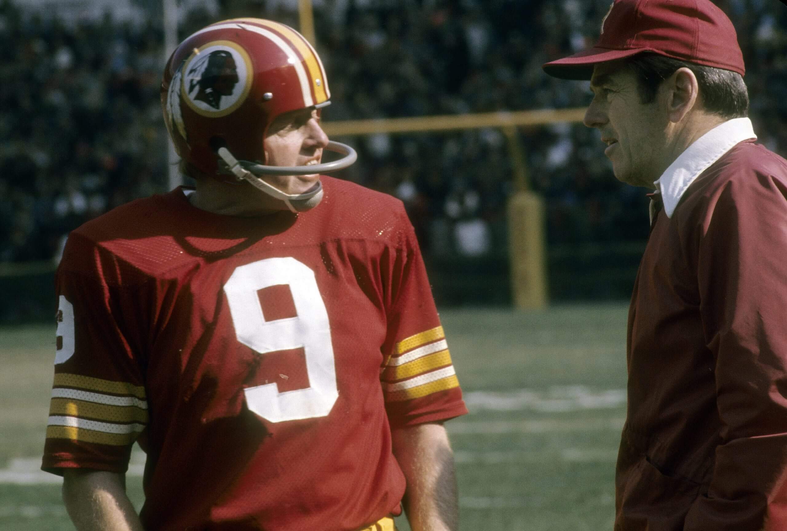 Sonny Jurgensen, on the left wearing a burgundy No. 9 uniform with a helmet with a single bar face mask, talks to coach George Allen, on the right wearing a burgundy hat and jacket with a white collar.
