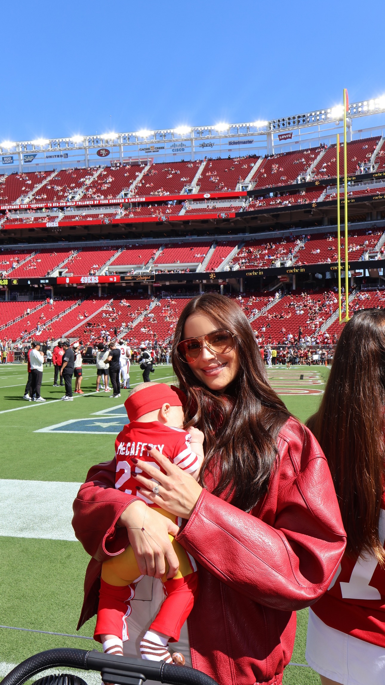 Olivia Culpo holding her daughter in a "McCaffrey" jersey at an NFL game.