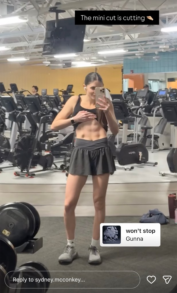 A woman in a sports bra and skirt in a gym lifts her shirt to show off her abdominal muscles.
