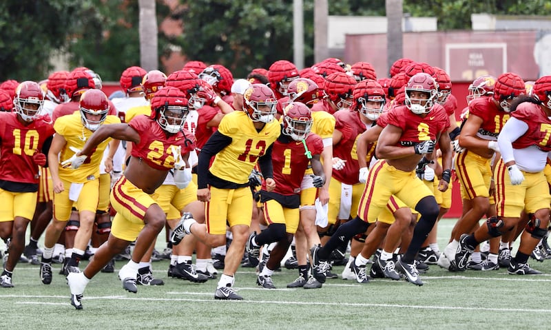A crowd of USC football players in cardinal and gold jerseys run up the practice field, led by Caleb Williams (13).