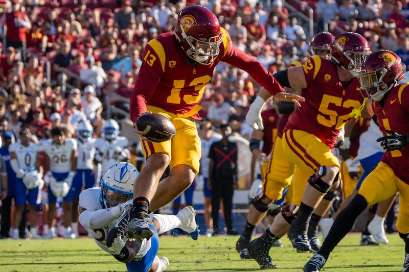 Williams (13) jumps with football in hand as an SJSU defender grabs at his ankles. Williams wears a cardinal and gold football uniform and the defender wears a white and blue football uniform.